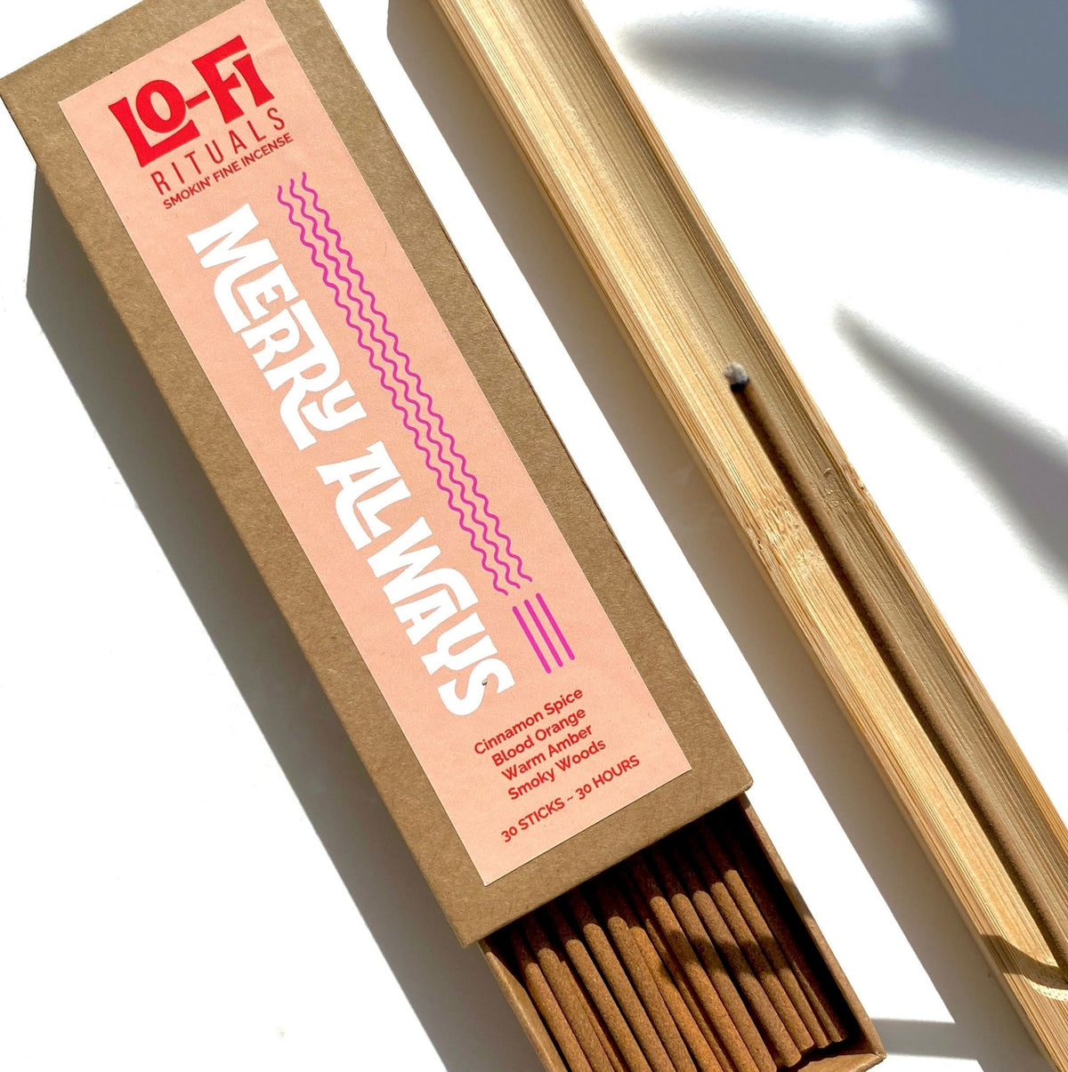 A box of Lo-Fi Rituals Merry Always incense, with it's bold and modern label is open, alongside a bamboo incense holder burning a stick of incense. They sit on a bright white background.