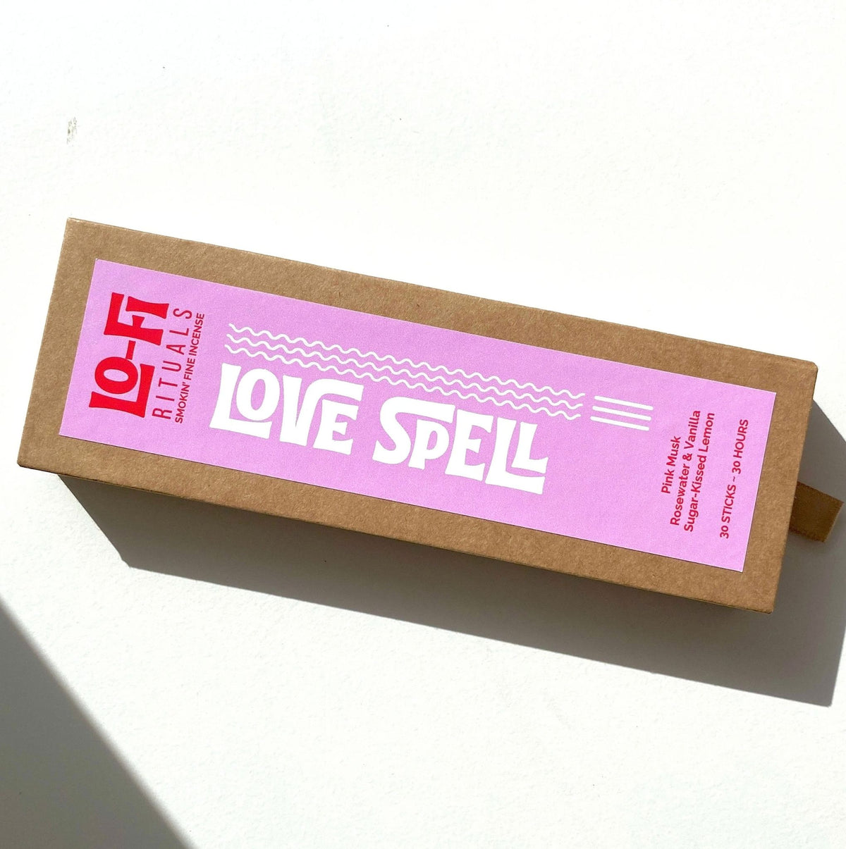 A rectangular brown kraft paper box with a pink label reading 'LO-FI RITUALS LOVE SPELL' in white text, photographed on a white surface with a hard shadow