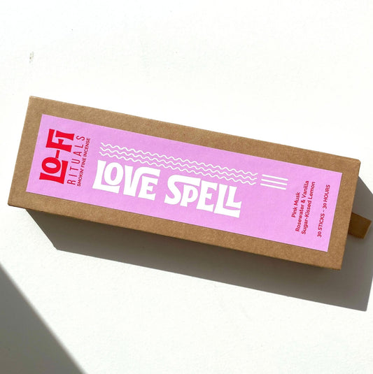 A rectangular brown kraft paper box with a pink label reading 'LO-FI RITUALS LOVE SPELL' in white text, photographed on a white surface with a hard shadow