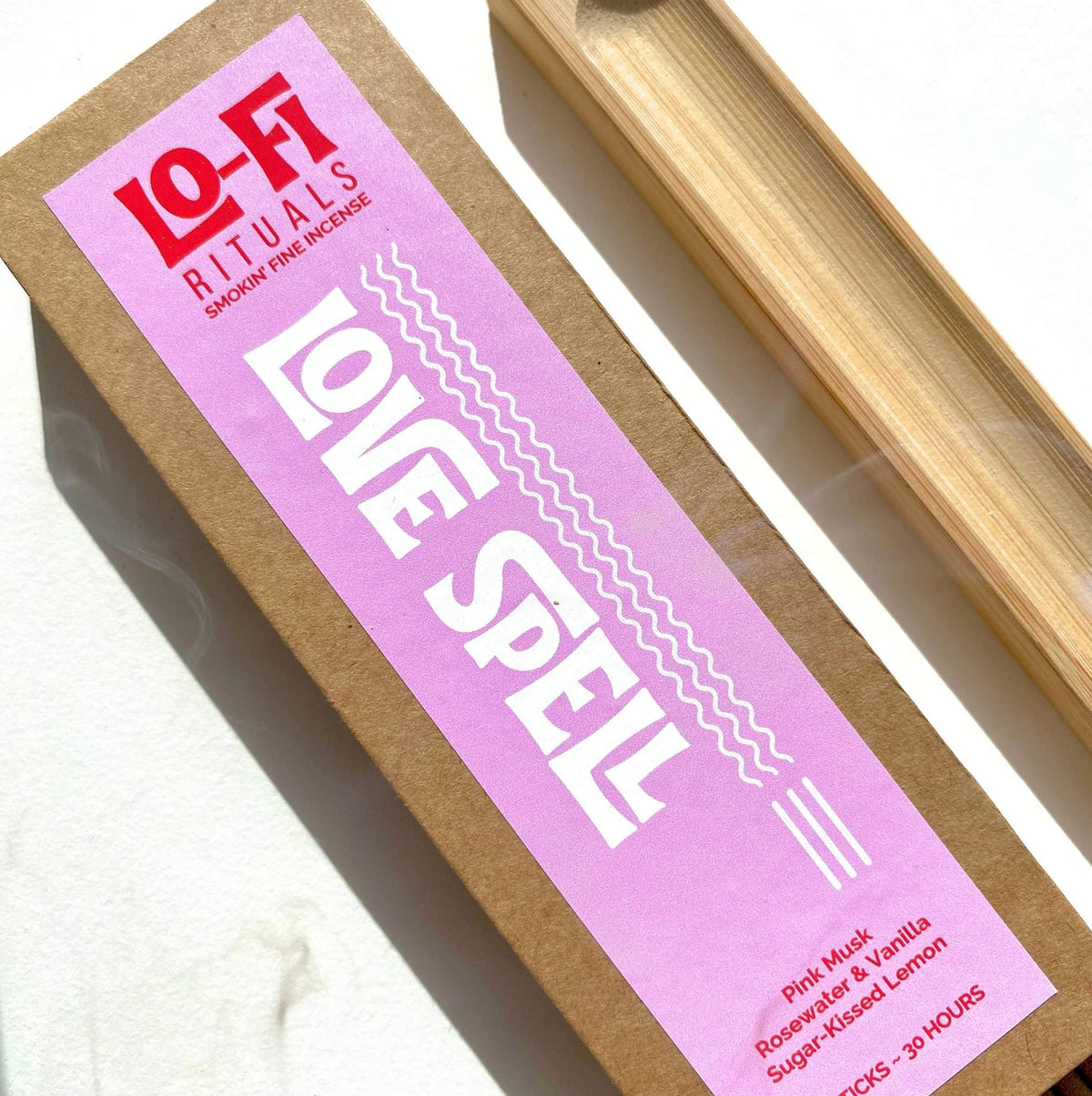 A kraft paper box of Lo-Fi Rituals Love Spell incense with a bright pink and red label, shown next to a bamboo incense holder on a sunlit white surface