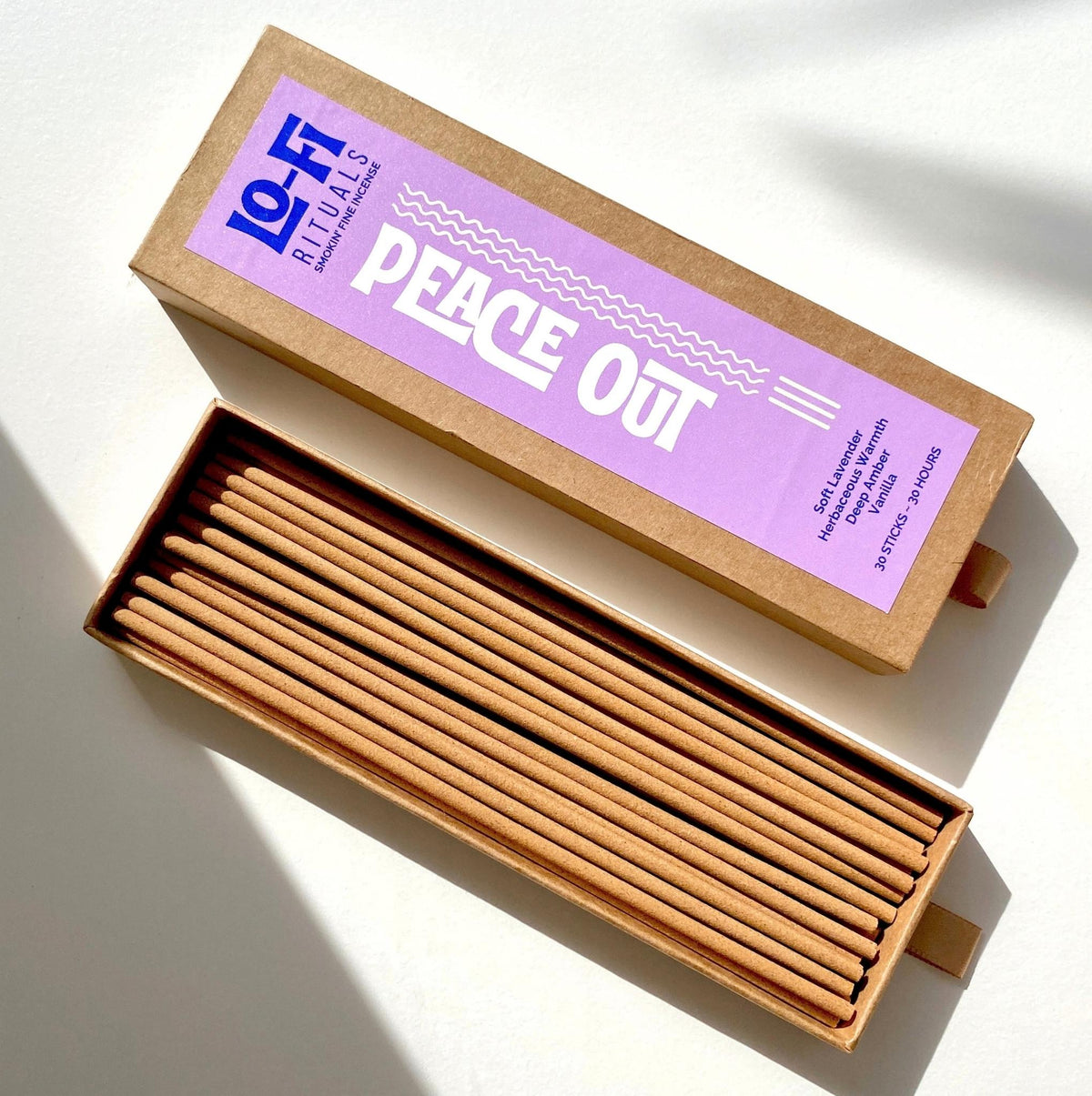 An open box of Lo-Fi Rituals Peace Out incense. The sun shines on the golden oak wood incense sticks. The modern, bold label includes scent notes for lavender, herbs, amber and vanilla.