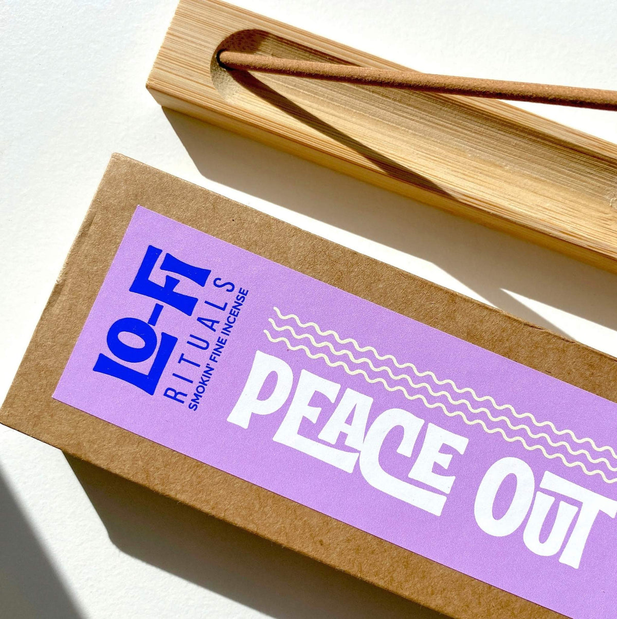 A close up image of Lo-Fi Rituals Peace Out incense, with bold lilac and blue label on a structured, quality kraft sliding drawer box. A bamboo holder sits beside with a lit incense stick.