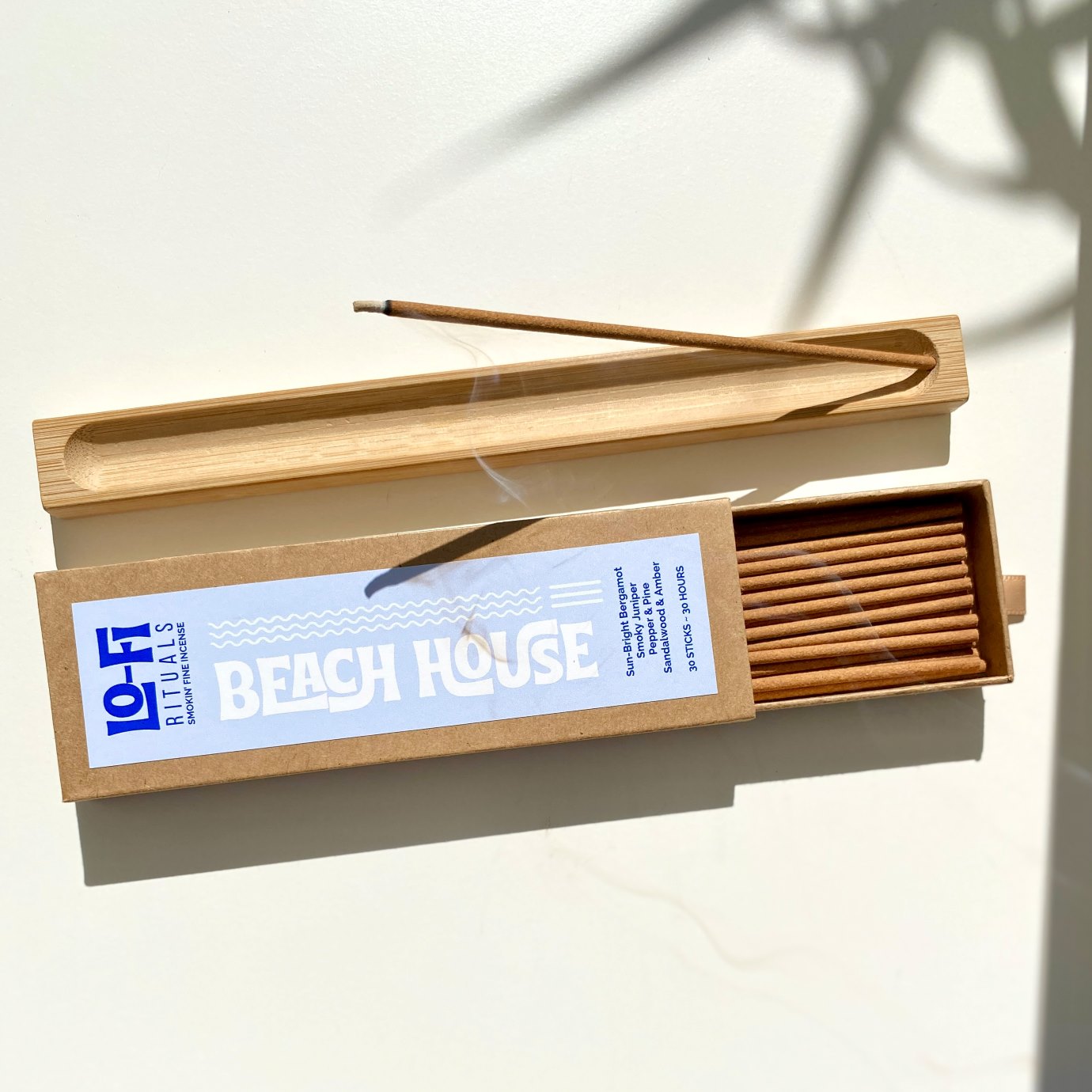 Burning incense in a wooden holder with curling smoke next to an open box of Lo-Fi Rituals Beach House incense sticks