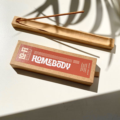 Styled image of Lo-Fi Rituals Homebody incense box, next to wooden incense holder on sunlit white background, framed by moody shadows