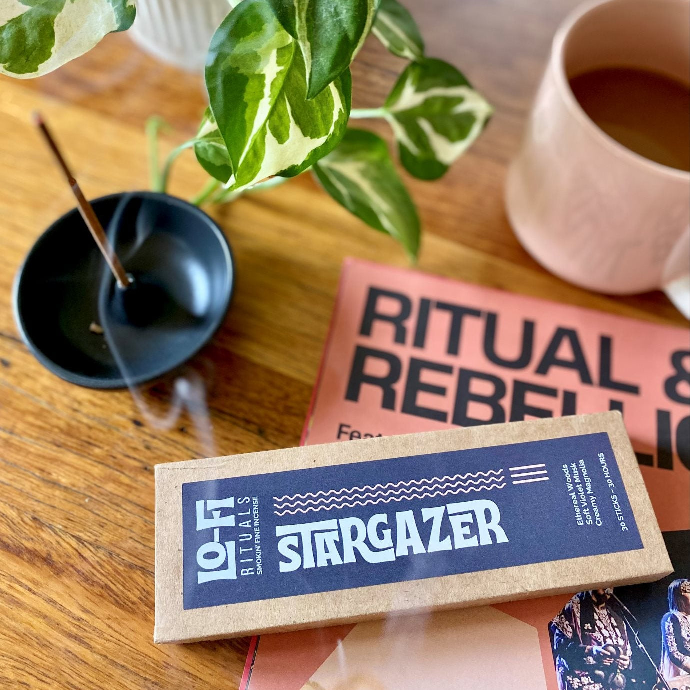 Lo-Fi Rituals Stargazer incense box on a table with a plant and mug in the background, incense burning in black ceramic holder with curling smoke
